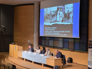 Author's photo of the second plenary with speakers sitting on the stage in front of a long desk, with a slide projected behind them